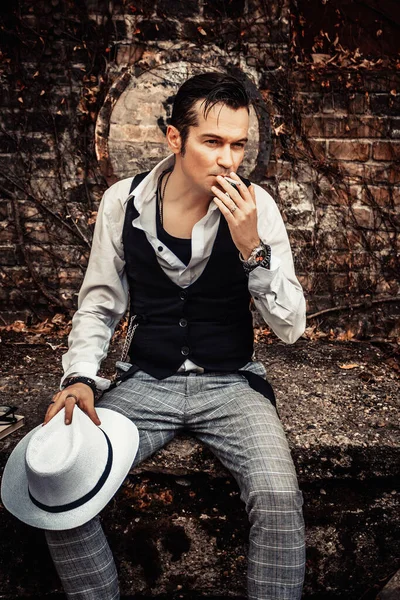Pensive man with fedora hat smoking cigarette while relaxing outdoors.