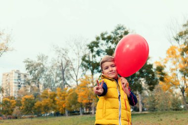 Cute kid with re balloon shoving victory sign during autumn day at the park.