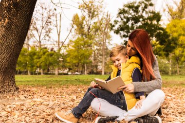 Small boy sitting on mother's lap while she is reading him book during autumn day in nature. 
