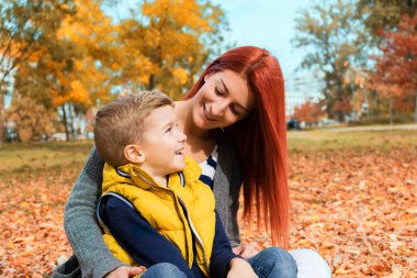 Happy mother talking with her small son while relaxing in autumn leaves and enjoying in the park. 
