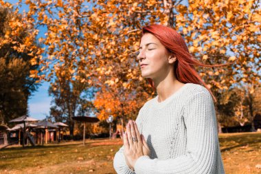 Redhead woman in namaste pose meditating in autumn day at the park. 