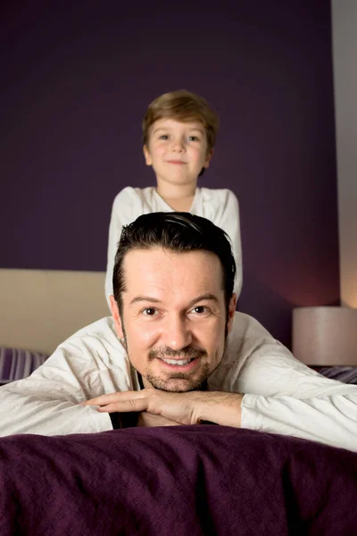 Happy father relaxing on bed while son is sitting on his back in the bedroom.