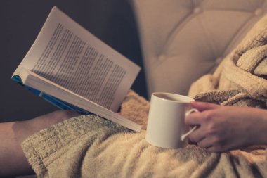 Close up of woman drinking tea and reading a book at night at home. 
