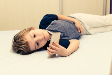 Small boy relaxing in the evening and watching cartoons on smart phone in bedroom. 