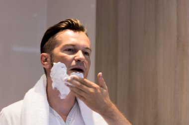 Reflection in a mirror of man applying shaving cream of face.