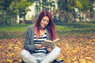 Young redhead girl reading book in autumn park. Happy red-haired woman studying and listening music over earphones in nature. 