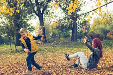 Happy boy and his mother playing and throwing leaves at each other in the park. Playful mother and son having fun in autumn park. 