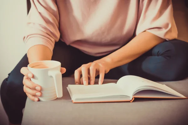 Unrecognizable woman drinking coffee and reading book while relaxing at home.