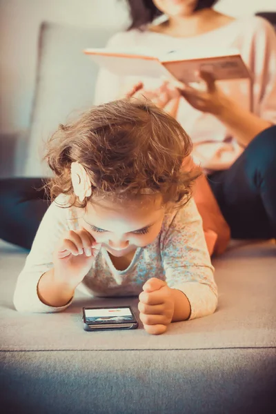 Cute small girl surfing the net on smart phone while relaxing at home with her mother.