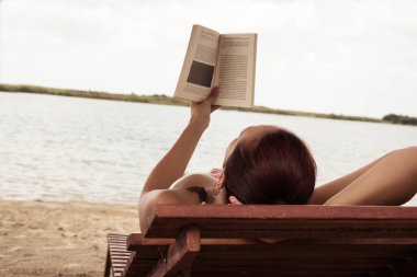 Sahilde güverte sandalyesinde uzanmış kitap okuyan tasasız bir kadın.. 