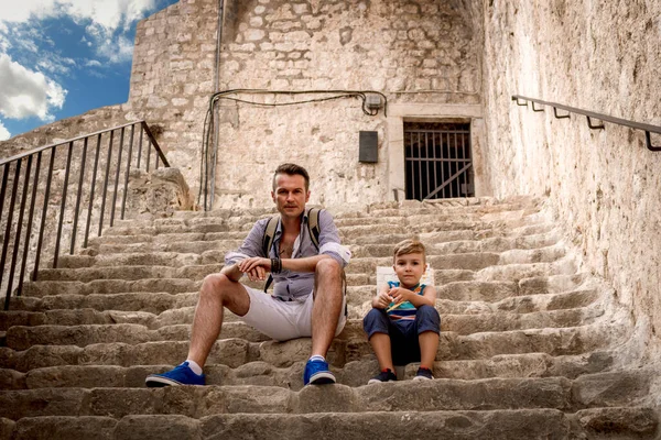 Father and son sitting on cobblestone stair while relaxing in old town during summer vacation.