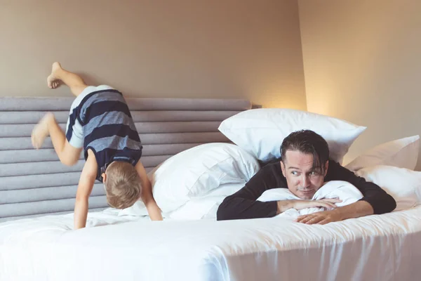 Father covered with pillow lying down on the bed while son is being mischief and climbing on the bed wall.