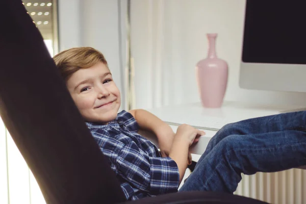 カメラを見ながら家でコンピュータを使用して笑顔の子供とテーブルの上に足で椅子でリラックス — ストック写真
