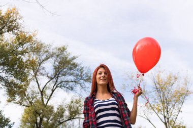 Parkta elinde kırmızı balonla gülümseyen kızıl saçlı kızın alçak açılı görüntüsü. 