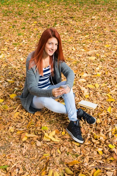 秋に座っている間にイヤフォンで音楽を聴く幸せな女性は自然の中で葉 — ストック写真