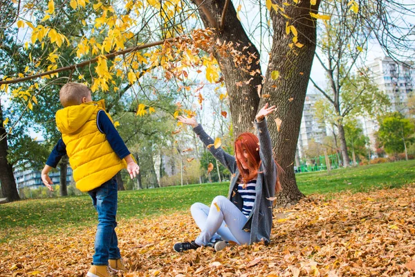 若い母親と彼女の息子投げ秋は空気中の葉と公園で楽しみを持っている — ストック写真