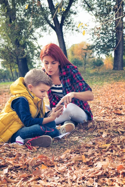 Küçük çocuk ve annesi sonbahar yapraklarında oturup internetten çizgi film izliyorlar. Genç anne oğluna parkta cep telefonu kullanmayı öğretiyor..