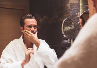 Bornozlu bir adam banyoda tıraş olduktan sonra nemlendirici sürüyor.. 