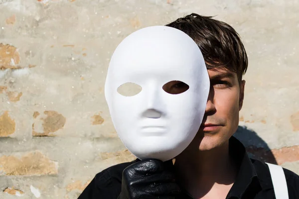 Man holding white mask over half of his face and looking at camera.