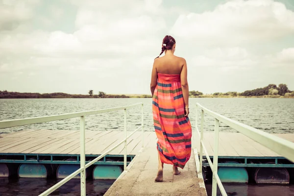 Back view of woman in sun dress walking on a pier at the beach. Copy space.