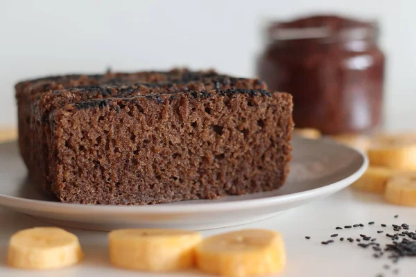 Ragi plantain cake. Gluten free tea cake made of Finger millet flour and pureed ripe plantain, sprinkled with black sesame seeds on top. Shot on white background with plantain slices and ragi flour around.