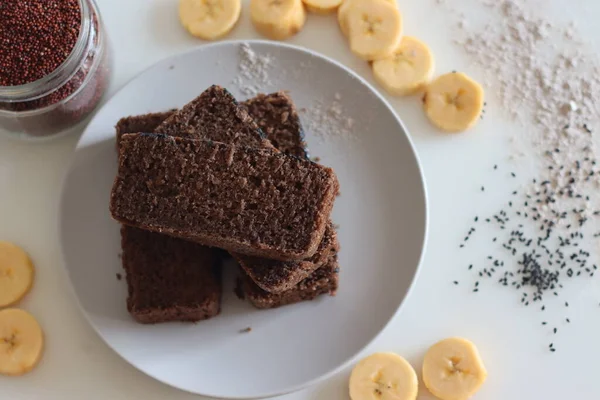 Ragi plantain cake. Gluten free tea cake made of Finger millet flour and pureed ripe plantain, sprinkled with black sesame seeds on top. Shot on white background with plantain slices and ragi flour around.