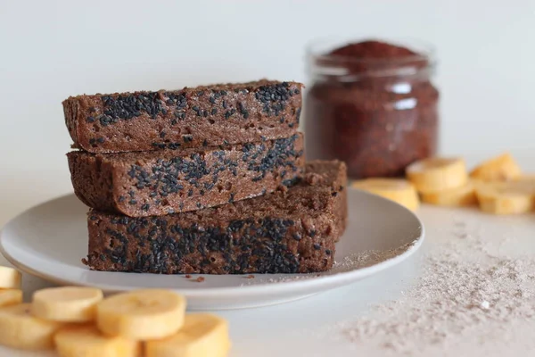 Ragi plantain cake. Gluten free tea cake made of Finger millet flour and pureed ripe plantain, sprinkled with black sesame seeds on top. Shot on white background with plantain slices and ragi flour around.