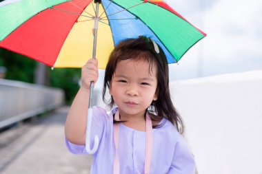 Anaokulu öğrencileri onun tenini doğrudan öğleden sonra güneş ışığından korumak için renkli şemsiyelerle yürürler. Tatlı bir kız eve yürüyor. Çocuk beyaz gömlek giyiyor..