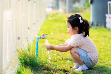 Şirin kız çömelmiş ve oyun parkındaki muslukta ellerini yıkıyor. Akşamları sıcak güneş ışığı. Bahar mevsiminde.