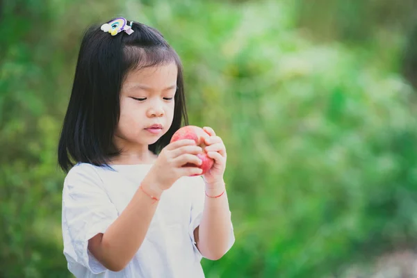 赤いりんごを持った可愛いアジア系の女の子 子供は空腹であり 新鮮な果物を食べる 子供たちは白いTシャツを着る 子供は４歳です 緑の自然背景 テキストを入力するための空のスペース — ストック写真