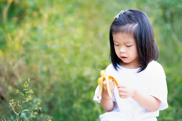 かわいい4歳のアジアの女の子は朝食のためにバナナを食べています 子供は黄色い果物を手に持っている バナナの皮むきの子供 自然食品 子供たちは白いTシャツを着る テキストを入力するための空のスペース — ストック写真