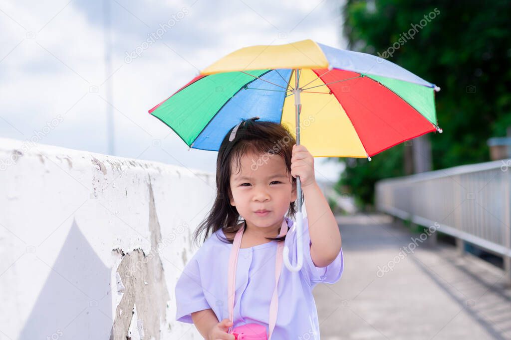 Portrait image of 4-5 years old. Cute Asian girl walks with umbrella in bright sun, to protect her from scorching sunny. In summer time. Kindergarten students are walking home after school. Copy space
