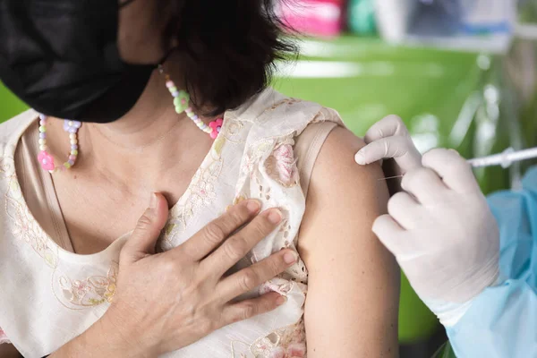 Siyah maske takan yaşlı bir kadının koluna aşı iğnesi tutan beyaz lastik eldivenli hemşire ya da doktorun eli, enfeksiyonu önlemek ve ciddi belirtiler göstermemek için..