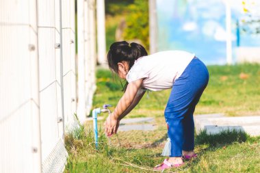 5-6 yaşlarında Asyalı bir çocuğun portresi. Küçük kız ellerini çimde ya da oyun alanında muslukta yıkıyordu. Öğleden sonra gün ışığı sıcakken. Yaz ya da bahar zamanı. Giriş alanı.