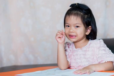 Ödevleri için çocuk benliği, ev eğitimi, sosyal uzaklık, e-öğrenim çevrimiçi eğitim, kalemle kız çizimi ya da yazısı, tatlı gülümsemeli mutlu çocuk, masada kitap, 4-5 yaş arası portre resmi çocuğu..