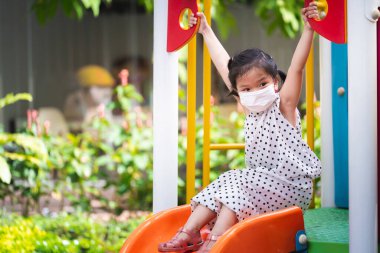Portre resmi 5-6 yaşlarında bir çocuk. Asyalı kız iki eliyle oyun parkında takılıyor. Çocuklar slayt oynar. Çocuk PM2.5 tozunu önlemek için yüz maskesi takıyor. Bebek dışarıda oyun oynuyor..
