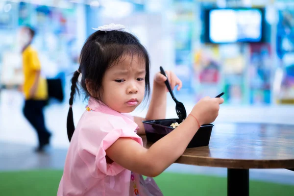 Asian cute girl is in bad mood while eating lunch or dinner.  Child frowned.  5 years old kindergarten kid is hungry.