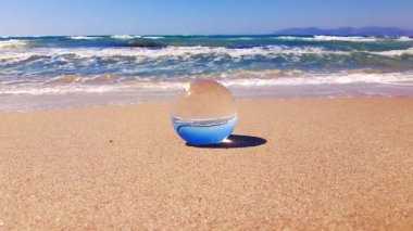 Hafif dalgalar, denizin önündeki kumda yatan cam topun etrafını yavaş çekimde siler..