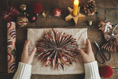 Kırsal ahşap masa üzerinde kırmızı kağıt yıldız ve süslemeli el sanatları kağıdında şık bir Noel hediyesi tutan eller. Atmosferik karamsar görüntü. Noel hediyelerini paketliyorum. Mutlu Noeller ve mutlu tatiller!