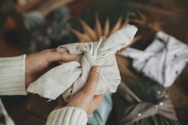 Furoshiki ambalajı, sıfır ziyan Noel tatili. Elleri, modern şenlik kumaşına sarılı noel hediyesini sade ahşap masa ve süslemelerle tutan eller. Atmosferik karamsar görüntü, kuzey tarzı