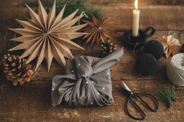 Kırsal ahşap masaya sarılı kahverengi kumaşla süslenmiş şık bir Noel hediyesi. Makas, kağıt yıldız, mum. Atmosferik karamsar görüntü, İskandinav tarzı. Furoshiki ambalajı, sıfır atık. Mutlu Noeller.!