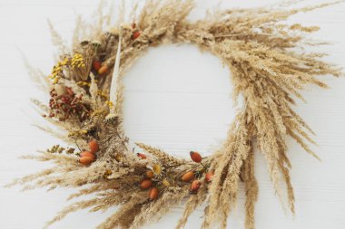 Şık bir sonbahar kır çelengi. Yaratıcı boho çelengi, kurutulmuş pampa otları, taneli kır çiçekleri, buğday, beyaz ahşap masa üzerinde köpek gülü meyveleri, izole. Mesaj için yer var. Sonbahar dekoru