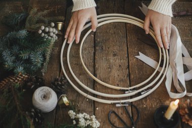 Eller, köknar dalları, esmer otlar, kurdeleler, tahta çember, makas, iplik, mum ile modern Noel çelengi yapıyor. Atmosferik karamsar görüntü. Kış tatili hazırlığı