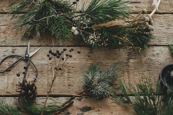 Modern christmas wreath with spruce branches, herbs, scissors, candle on rustic wood. Merry Christmas! Stylish boho xmas wreath details. Moody image, winter holiday preparation