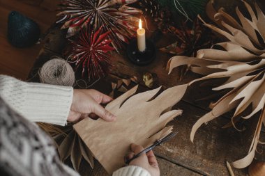 Şık Noel yıldızları yapmak. El yapımı kağıt torbadan yıldız kesen eller, arka planda İsveç yıldızları, iplik, mum, kırsal kesimde makas. Festivalsel dekor oluşturma süreci