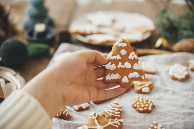 Elinde süslü zencefilli kurabiye Noel ağacı peçeteli, mumlu ve dekorasyonlu köy masasının arka planında. Huysuz bir görüntü. Tarz sahibi bir kadın Noel kurabiyesi yapıyor.