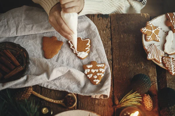 Hands decorating gingerbread cookie christmas tree with icing on rustic table with napkin, spices, candle, decorations. Top view. Moody image. Woman making stylish christmas gingerbread cookies