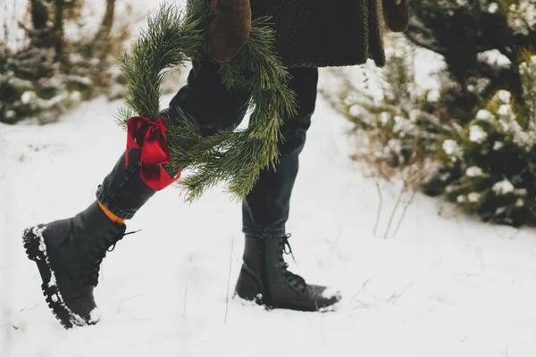 メリークリスマス 冬の公園でクリスマスリースとスタイリッシュな女性 若い女性で居心地の良いミトンとコート歩くとXmas花輪とともに赤い弓で雪の冬のフィールド クロップドビュー — ストック写真