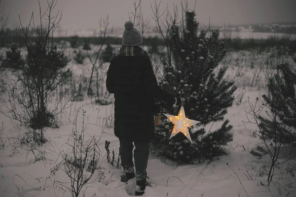 一个有着闪亮的大圣诞星的女人晚上在雪地的冬日公园里散步 神奇的冬季时间 在圣诞节前夕 时髦的嬉皮士女人在寻找奇迹 后视镜圣诞快乐 — 图库照片