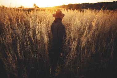 Şapkalı şık bir kadın sonbahar tarlasında çimlerin arasında sıcak günbatımının tadını çıkarıyor. Sakin atmosferik bir an. Akşamları kırsalda dikilen, eski moda elbiseli, şık genç bir kadın.